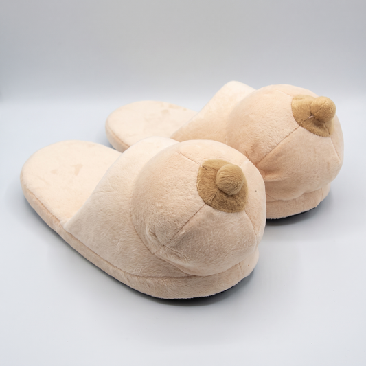 A pair of plush novelty woman's breasts slippers, in a soft beige color with a small brown accent, photographed on a light gray studio background.